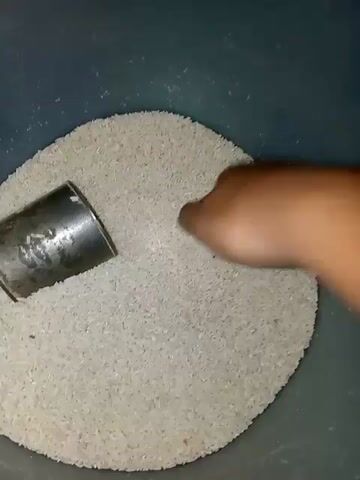 The video shows a close-up of a person's hand scooping a pile of uncooked white rice from a large, dark grey container. The hand is dark-skinned and appears to be an adult's. The rice is being scooped with a small, metallic tin. The container is mostly filled with rice, and the lighting is dim. The action is captured from a top-down perspective. The video is very short, only a few seconds long, and the camera angle and focus remain constant throughout.