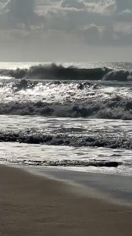 The video shows a wide shot of a beach with waves crashing on the shore. The sun is shining, creating a bright glare on the water and the wet sand. The waves are moderate in size, with white foam crests as they break. The sky is partly cloudy. The camera is static and positioned low to the ground, giving a ground-level perspective of the beach and ocean. The scene is natural and serene.