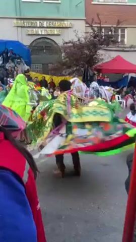The video shows a street parade of people wearing large, bright, colorful, patterned outfits. They are dancing and spinning around in the street. The buildings behind them are light green and red brick, with windows. There is a crowd of people watching the parade on the sidelines, sitting in chairs. A red tent is also visible in the background. The parade appears to be taking place on a paved street in an urban environment.