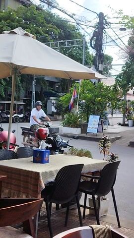 This media file captures an outdoor scene in Kampot, Cambodia. The primary subject is a man wearing a white t-shirt and a helmet, sitting on a motorcycle. He is positioned near a table with two black chairs and a beige tablecloth, suggesting a cafe or restaurant setting. A white umbrella, partially open, is set up nearby, providing shade.

The background reveals a street scene with lush greenery and buildings. Several motorcycles are visible, indicating a common mode of transportation in the area. A flag with red and blue colors is also present, possibly a national or regional flag. The architecture suggests a tropical climate, with open spaces and vegetation. The time of day appears to be daytime, with bright lighting and shadows.

Notable details include a red helmet on the ground next to the motorcycle and a blue box on the table. The overall atmosphere seems relaxed and casual, typical of a street-side establishment. The man on the motorcycle is looking towards the left of the frame, his expression not clearly discernible. The presence of electrical wires and poles in the background are typical of urban infrastructure in many parts of Southeast Asia. The scene is characterized by a blend of urban elements and natural tropical flora.