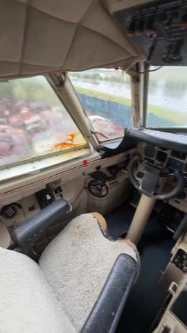 The camera pans around the cockpit of a plane on a rainy day. The cockpit is tan and black with worn seats. Various gauges and controls are visible, and the windscreen has water droplets on it. The plane appears to be stationary.