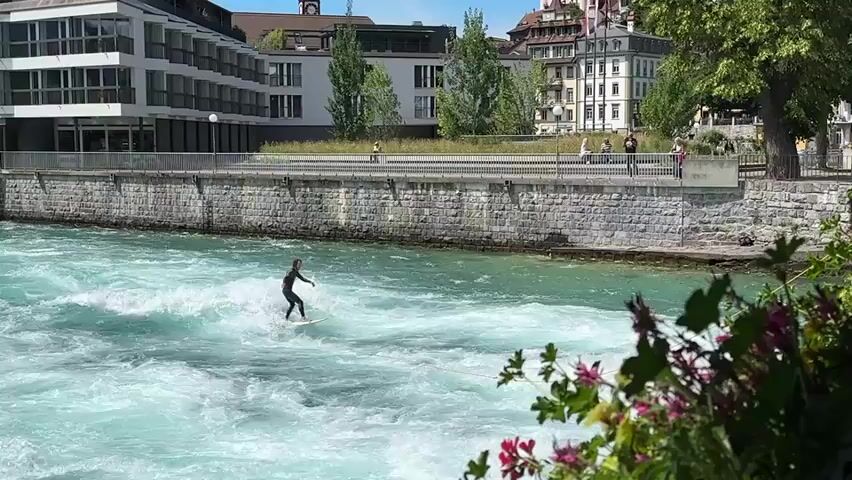 A person wearing a black wetsuit is surfing on a white surfboard in a river with turbulent, turquoise water. The surfer is positioned to the left of the frame and is being pulled by a yellow rope, indicating they are being towed. The riverbank on the right is lined with lush green foliage, with some pink and purple flowers in the foreground. On the left side of the river, a modern building with a glass facade stands beside a stone wall. Further back, several older buildings with traditional architecture are visible. A metal fence separates the riverbank from a walkway where a few people are strolling. The sky is clear and blue.