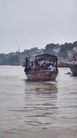 A boat carrying several people wearing orange life vests travels on a muddy river. The boat has a covered roof and appears to be moving away from the camera. In the background, a city with numerous buildings lines the riverbank. The sky is overcast.
