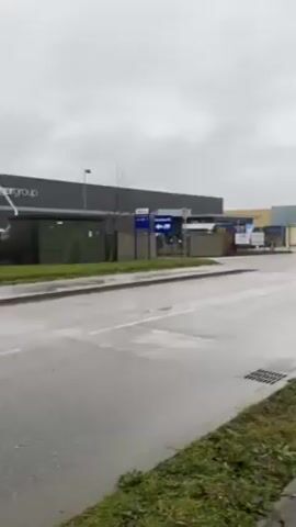 The video shows a white and blue truck with "loginte" on the side driving on a wet, gray road. The truck is pulling an empty car carrier trailer. A yellow sign with "SORTIE DE CAMIONS" is visible on the right side of the road. There are industrial buildings in the background under a cloudy sky.