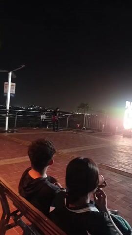 The video shows a nighttime scene at a public promenade. In the foreground, two people are sitting on a bench facing away from the camera. The person on the left has dark curly hair, and the person on the right has dark hair styled in a ponytail. The person on the right appears to be holding a drink and smoking or holding a cigarette. The promenade is paved with brickwork, and a large illuminated sign that reads "PLAZA JOSÉ JOAQUÍN DE OLMEDO GUAYAQUIL" is visible in the background. Several other people are walking around in the area, and there is a street performer playing a guitar in the distance. The environment is dimly lit by streetlights and the glowing sign, with the sky appearing dark and starry.