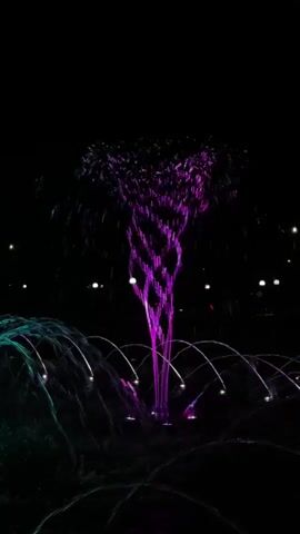 A water fountain is illuminated with colored lights in the dark. The fountain shoots water upwards in a twisting column that changes color from purple to blue to green. Smaller streams of water arc around the base of the fountain. The background is dark with a few blurred lights visible, suggesting it is nighttime and the fountain is outdoors.