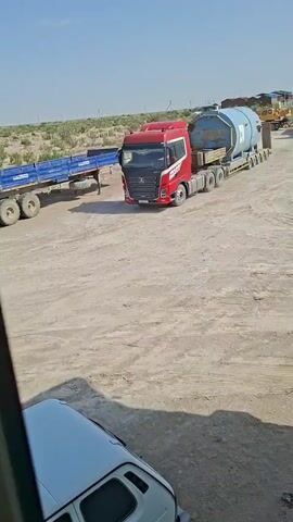 A red and black semi-truck is transporting a large blue cylindrical object on a flatbed trailer. The truck is driving on a dirt road, with other vehicles and construction equipment visible in the background. The sky is clear and blue, and the sun is shining.
