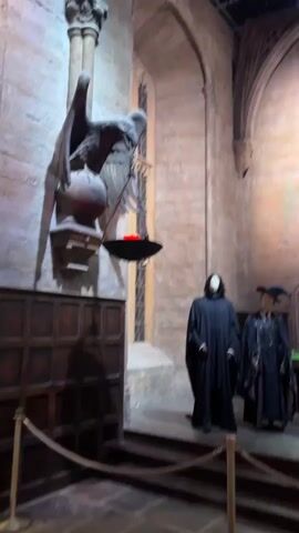 This video captures a scene from the Great Hall at Warner Bros. Studio Tour London – The Making of Harry Potter. The scene depicts the enchanted ceiling of the Great Hall, a hallmark of the Harry Potter film series. Various food items are laid out on long wooden tables, suggesting a feast or a meal is taking place. Two mannequins are dressed in dark robes and wizarding attire, positioned to represent characters from the films. One mannequin, with a pale, smooth face and long dark hair, is likely intended to represent Professor Severus Snape. The other, wearing a pointed hat and dark robes, appears to be a witch. Further down the hall, another mannequin, adorned in elaborate robes and sporting a long white beard, is dressed as Albus Dumbledore, the headmaster of Hogwarts. The setting is grand and atmospheric, with high arched ceilings, stone walls, and large, ornate windows that suggest a medieval or gothic architectural style. The lighting is dim, creating a sense of mystery and enchantment, typical of the Hogwarts Great Hall. The video appears to be filmed during the daytime, given the artificial lighting in the studio setting. There is no discernible text visible in the video. The overall impression is one of immersion into the magical world of Harry Potter, showcasing a meticulously recreated film set.