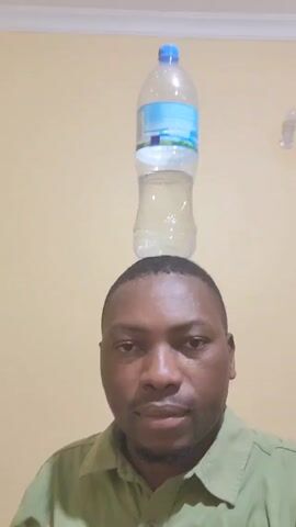 The video shows a man in a light green shirt with a short haircut looking directly at the camera. He is indoors and a water bottle with a blue cap is balanced on his head. The wall behind him is a light yellowish color.