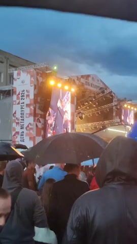 The video shows a crowded outdoor event with a stage in the background. A performer is visible on the stage, which is lit up with bright lights and smoke effects. The crowd is gathered in front of the stage, many holding umbrellas due to what appears to be rain. The overall atmosphere suggests a concert or festival. The stage backdrop features a banner with text that includes "ДЕНЬ МЕТАЛЛУРГА 2026", indicating the event is a celebration for metallurgists in 2026. People are dressed in casual clothing, and some are wearing hoods, further suggesting inclement weather. The lighting is dim, consistent with an evening or overcast sky.