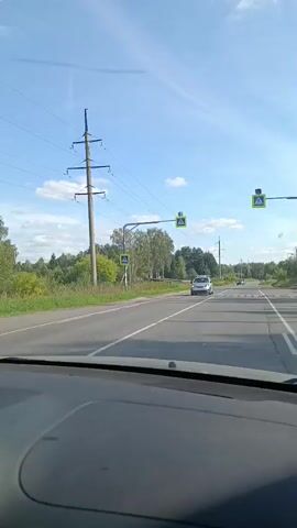 The video shows a view from inside a car driving on a two-lane road. The road is paved and has white lane markings. There are trees and vegetation on both sides of the road, and power lines are visible overhead. Several cars are also seen on the road. The sky is blue with some clouds. There are pedestrian crossing signs with flashing lights at intervals along the road. The overall environment appears to be rural or suburban.