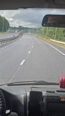The video is from the perspective of someone driving a vehicle. The road ahead is wet, indicating it may have recently rained. There are lane markings on the road, and guardrails line the left side. Trees and poles with lights are visible along the sides of the road. In the distance, other vehicles can be seen. The dashboard of the vehicle is visible in the foreground, with a phone mounted on a holder. A hand is on the steering wheel. The sky is cloudy.