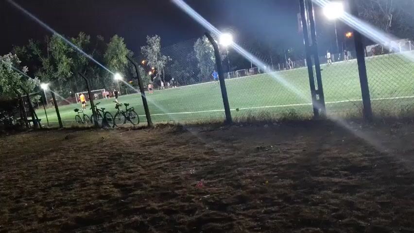 The scene is at night on a soccer field enclosed by a chain-link fence. Three bicycles are parked along the fence in the foreground. Players are on the field, lit by bright stadium lights, engaging in what appears to be a soccer game or practice. Trees line the perimeter of the field in the background. The ground in the foreground is covered with dry grass and dirt.