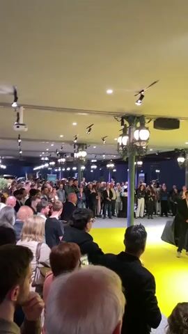 The video depicts an indoor event, likely a product launch or presentation, taking place in Mulhouse, France. The scene features a brightly lit stage with a vibrant yellow floor covering the entire visible area, where an audience is gathered, both seated in the foreground and standing further back.

On the stage, several individuals are present. A man in a dark suit is speaking into a microphone, addressing the attendees. To his left, another man in a dark suit stands near a car that is completely draped in a black sheet, suggesting an upcoming unveiling. To the right of the speaker, three other individuals – two men and a woman – stand facing the audience.

The backdrop is a large yellow wall featuring prominent white text and a logo. The central element is a large "ipop" logo. To the right of this, text in French reads "LA NOUVELLE VOITURE POPULAIRE" (The new popular car), followed by "4 PLACES," "PENSEE ET FABRIQUE EN FRANCE" (Conceived and manufactured in France), and "EDF." On the left side of the backdrop, smaller, partially visible text includes "28 AVRIL" and "MULHOUSE," providing further context to the event's location and date.

The venue's ceiling is equipped with multiple spotlights and decorative chandeliers, illuminating the space. A man operating a video camera can be seen near the covered car, recording the event. The atmosphere appears formal and anticipatory, typical of a launch event for a new vehicle.