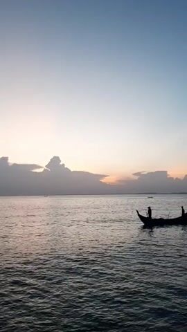 Here's a description of the video:

The video shows a boat moving on the water at sunset. There are two people visible in the boat. The sky is a mix of light blue and orange hues, with clouds obscuring the sun on the horizon. The water is slightly choppy. There are other small boats visible in the distance. The boat in the foreground is silhouetted against the bright sky.