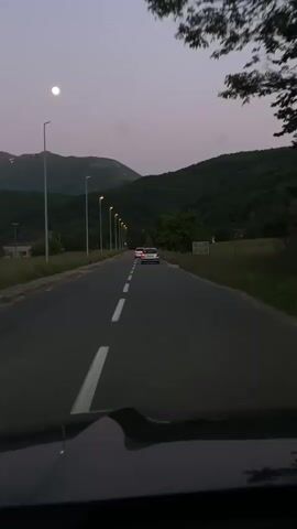 The video depicts a car journey on a road at dusk. The sky is a dusky pink, with a full moon visible. The road is asphalt with white lane markings. There are streetlights illuminating the road. In the distance, there are mountains and green hills. Two cars are visible ahead on the road. The scene is filmed from the perspective of the driver inside a car, showing the dashboard.