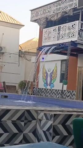 The scene showcases a swimming pool with water flowing into it from a decorative water feature. The water feature consists of multiple streams cascading down from the upper story of a building. The building has ornate, white latticework on its balcony and a mural of rainbow-colored wings with a crown above them on the wall beside the pool. The pool is bordered by a tile pattern of black and white chevrons. Other buildings are visible in the background, along with some greenery and other minor objects.