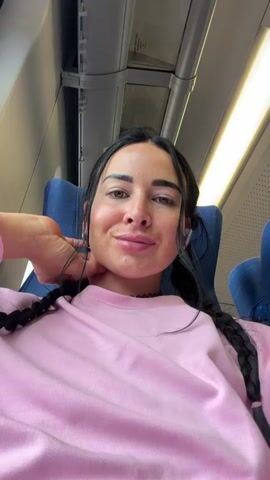 A woman with dark hair in braids, wearing a pink sweatshirt, is sitting in a train seat. She is looking at the camera and touching her neck with her left hand. She has white earbuds in her ears. The interior of the train is visible behind her, with blue seats and overhead compartments. The lighting appears to be artificial.