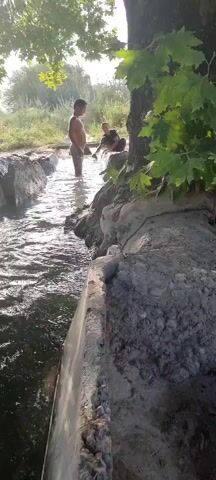 The video shows a natural irrigation ditch flowing through a rural area. Two young people, a girl and a boy, are visible in the water, suggesting they are playing or cooling off. The girl stands near the middle of the ditch, and the boy is further downstream. The scene is set under the shade of a large tree with broad leaves, indicating it's likely a warm day, possibly late morning or afternoon due to the sunlight filtering through the leaves. The surrounding landscape includes dry, sandy ground and lush green fields in the background, suggesting an agricultural area. The overall mood appears relaxed and playful, with children enjoying the water in a natural setting. There is no visible text in the video.