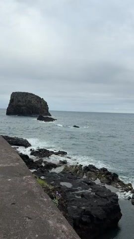 The video shows a view of the ocean coastline from a slightly elevated position, likely a concrete walkway or wall. The sky is overcast. In the distance, there's a rocky island or a large rock formation in the ocean, along with glimpses of land with green hills. 
The foreground features dark, jagged rocks along the shoreline, and waves are crashing against them. There is a set of stone steps leading down to the rocky area. The water is a mix of dark and light blue due to the light and the waves.