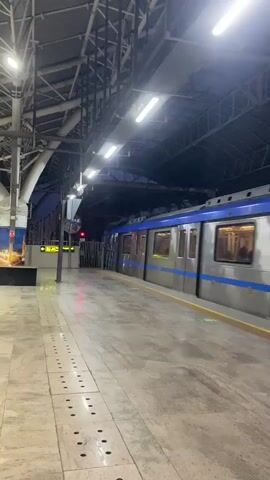 This media file captures a view of a metro train at a station. The train is silver with a blue stripe and is positioned at the platform. The station appears to be indoors, with a large, industrial-style roof structure supported by metal beams. Fluorescent lights illuminate the platform and the train. The platform itself is paved with large, light-colored tiles, and a tactile paving strip is visible along the edge, indicating it's designed for visually impaired passengers.

To the left of the frame, there's a large advertisement or mural depicting a city skyline with a bright, colorful sunset. Further down the platform, a yellow directional sign with text is partially visible. A red traffic light or indicator is also present near the sign. The overall lighting suggests it might be late evening or early morning, or that the station is artificially lit. The scene is quiet and still, with no people visible on the platform or inside the train, creating a sense of solitude or anticipation. The metallic structure of the station roof and the sleek design of the train give a modern and functional impression. The location is identified as St.Thomas Mount-Pallavaram Cantonment Board, India.