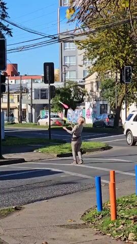 A person juggles three batons in a crosswalk. The traffic signal shows red. The person completes the juggling and walks off camera. Traffic passes.