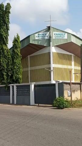 The video shows a view of the Holy Catholic Family Church Ajara Badagry in Nigeria. The church is a one-story building with a distinctive architectural design featuring vertical yellow stripes and a light green roof. A cross is mounted on the roof. The church is enclosed by a gray and white fence. Several tall, thin green trees stand beside the church, adding to the building's height. The video also shows a motorcycle carrying three people driving past the church. The environment appears to be a suburban street with a clear sky and a few scattered clouds. The video is taken from inside a moving vehicle.