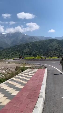 A woman in a green jumpsuit walks on a paved road next to a sidewalk. In the background, there are mountains covered with green trees and a cloudy blue sky. Further in the distance, there are people on a long suspension bridge, and a roller coaster track. The video pans from left to right, following the woman and the landscape.
