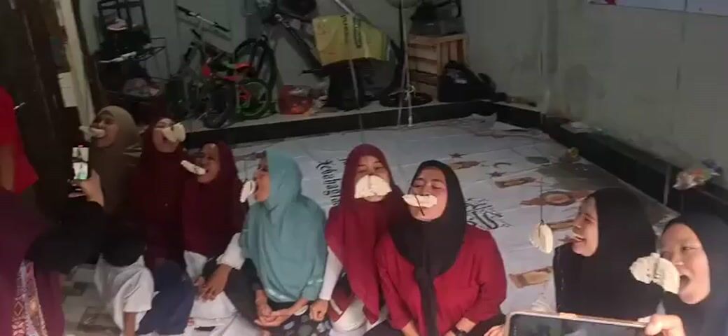 The video shows a group of at least seven women sitting on the floor, likely indoors. Each woman is wearing a hijab. Several of them have white objects, possibly crackers or snacks, attached to strings hanging from above, which they are attempting to eat without using their hands. One woman on the far left is holding a smartphone, presumably recording or taking pictures. The environment appears to be a room with a tiled floor and a dark wall in the background. There are some bicycles and boxes visible in the background, suggesting a somewhat cluttered or utilitarian space. The women appear to be participating in a game or competition.