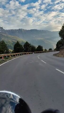 A first-person perspective video of someone riding a motorcycle or similar vehicle on a winding mountain road. The road is paved and has white dashed lines marking the lanes. A guardrail with yellow and black stripes is visible on the left side of the road. The surrounding environment is mountainous with lush green trees and a vast valley. The sky is partly cloudy with the sun peeking through, casting light and shadows on the landscape. The video captures the journey along the scenic route.