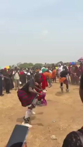 The video captures a traditional cultural dance performance in an outdoor setting, likely a celebration or festival. Several individuals dressed in colorful, traditional costumes are dancing in the center of a crowd of people. The dancers are wearing costumes featuring red, green, and black patterns. One dancer wears a large green hat and carries orange pom-poms. The crowd includes people of various ages and attires. The ground is dry and dusty. A person in the foreground is filming the scene with a smartphone. Baskets of produce are carried by some in the crowd.