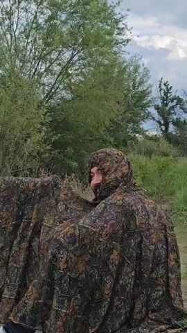 A person in a camouflage poncho is holding a camera with a long lens next to a river. The camera is mounted on a tripod. The person is looking toward the river, which has vegetation growing along the shore. The sky is cloudy.