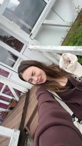 A young woman with long brown hair and a brown hoodie is taking a selfie. She is standing outside next to a wooden structure with white railings. In the background, there is a red wooden house with a white porch, trees, and a blue inflatable pool. The sky is cloudy.