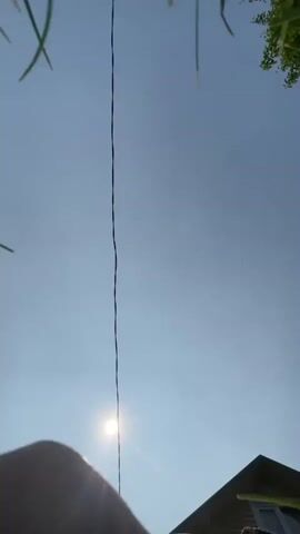 A person wearing a hat and a long-sleeved shirt walks into the frame from the bottom of the screen. The person is walking towards a thin, dark, vertical line that extends from the top of the screen to the bottom. The sun is visible behind the line, and a house is partially visible in the bottom right corner of the frame. The sky is clear and blue.