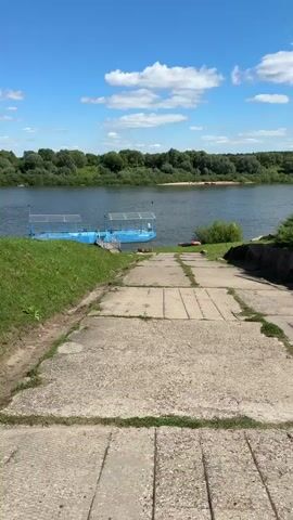 A slightly elevated, wide concrete ramp slopes down toward a wide, calm river. The ramp's surface is uneven, with visible cracks and debris. To the left of the ramp, a steep, grassy embankment rises. On the river, a blue, rectangular pontoon is docked. It has a blue railing and a light blue roof supported by metal poles. A wooden walkway leads from the shore to the pontoon. On the far bank of the river, a dense line of green trees runs along the horizon. The sky is bright blue with scattered white clouds. In the foreground, on the right side of the ramp, is a hint of a red object, possibly a float or a piece of debris. The scene appears to be a public access point to the river on a sunny day.