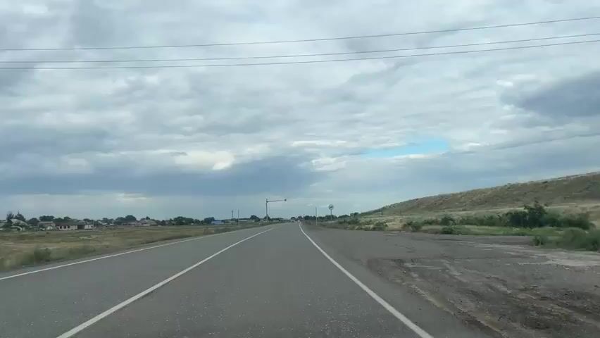A wide asphalt road with two white lines marking the lanes stretches towards the horizon under a cloudy sky. On the left side of the road, a village with houses is visible in the distance. On the right side, a grassy embankment slopes upwards, dotted with dry vegetation. Traffic signals and signs are present along the roadside. The camera is positioned inside a vehicle, looking forward.
