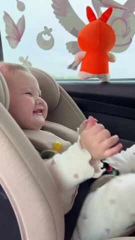 A happy baby is securely fastened into a light beige car seat inside a vehicle. The infant is wearing a white long-sleeved top with ruffled cuffs and white pants adorned with small heart patterns. A yellow pacifier is attached to its clothing with a pink and white beaded clip.

Throughout the video, the baby consistently displays a wide, joyful smile, often looking directly towards the camera and occasionally moving its hands.

The car's interior is visible, and the window features several decorative decals, including a pink butterfly, a white baby bib, a white baby pacifier, and a white winged creature, possibly a bird or angel. An orange plush toy with prominent ears also hangs near the window.

Through the car window, a cloudy sky and green fields or trees indicate that the car is in motion during the daytime, possibly traveling through a rural or semi-rural area consistent with Staroderevyankovskaya, Russia. Upbeat music can be heard playing in the background. No visible text is present in the video.
