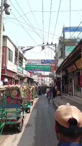 The video shows a narrow street in what appears to be a bustling urban environment. The street is lined with buildings, many of which appear to be shops with signs in a language other than English. Overhead, there are numerous banners and electrical wires crisscrossing the sky. Several auto-rickshaws are parked along the left side of the street, their colorful designs standing out. People are walking in both directions along the street. The camera appears to be handheld, positioned behind and slightly to the side of someone wearing a tan baseball cap. The overall impression is one of a busy, vibrant market or shopping district in a developing country.