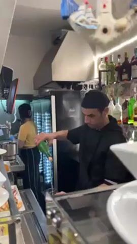 Here is a concise description of the video. 

**Setting:** The video appears to be taken in a bar or restaurant kitchen. 

**Subjects and Objects:** A man in a black chef's jacket and hat is pouring a green bottle of liquid into a glass. There is a woman in the background in a yellow shirt and dark pants. Various food items are visible in dishes on the counter, including some with oil. Shelves behind the counter are stocked with bottles of alcohol. There is a refrigerator and a kitchen hood in the background.

**Action:** The man is carefully pouring the liquid from the green bottle into a glass. The woman in the background appears to be working.