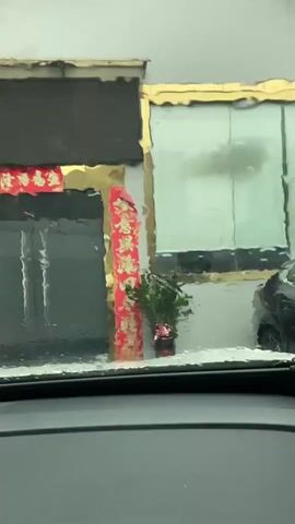 The video, filmed from inside a car, shows a store entrance decorated with red banners with gold lettering. The banners are hung vertically on either side of the entrance and horizontally above it. A small potted plant with green leaves and pink flowers is placed to the right of the entrance. The entrance itself appears to be a set of double doors. The view is slightly distorted by rain on the car's windshield. The interior of the car, including part of the dashboard and a rear-view mirror, is visible in the foreground. The exterior scene is somewhat blurry and appears to be a street or alleyway.