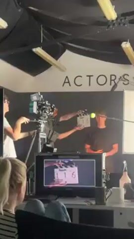 Here's a description of the video:
A film shoot is taking place indoors. A young man stands center frame, facing the camera with a tennis ball seemingly stuck to his forehead. A video camera on a tripod is positioned to his left, operated by someone out of frame. Another person holds a clapperboard near the camera. A video monitor in the foreground displays the live feed from the camera, showing the young man’s face. To the left of the monitor, a person with blonde hair sits and watches the monitor. The background wall is black with the word "ACTORS" displayed in large white letters. Fluorescent lights are mounted on the ceiling. Other items like cups, a spray bottle, and a whiteboard are visible on a table near the monitor.