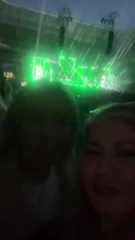 A selfie video taken at a concert. Two women are in the foreground, smiling. They are in a crowded stadium with a stage and bright green lights in the background. The concert appears to be underway.