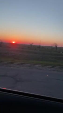 The video shows a car driving on a rural road at sunset. The sun is a bright red ball on the horizon.  A field and a large, white, barn-like building are visible.  The road is cracked and patched. The video then switches to a shot over the steering wheel, showing the road ahead and another car in the distance.  The dashboard gauges are visible, including the speedometer. The sky is a clear gradient of blue and orange.