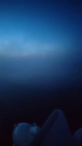 The video shows a body of water, likely a lake or river, at dawn or dusk.  A long concrete pier or breakwater extends into the water.  The sky is a gradient of dark blue to a lighter orange/pink on the horizon.  A bank of fog obscures part of the distant shoreline and the water surface, leaving only a thin horizontal reflection visible.  The water close to the pier shows ripples, indicating movement, either from wind or possibly a passing boat. The camera appears to be moving forward.