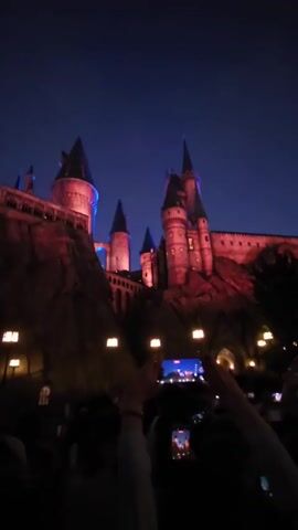 The video shows a crowd of people gathered at night, holding up their smartphones to film a castle-like structure. The castle is illuminated with red and blue lights, creating a dramatic effect against the dark blue night sky. The architecture suggests a fantasy or medieval theme, possibly a replica of a famous landmark. The people are silhouetted against the lights and the castle, with their faces largely obscured. Their raised phones indicate they are capturing a performance or display. The atmosphere appears to be one of excitement and anticipation, common for nighttime attractions or events. The presence of multiple smartphones suggests a public event where people are eager to document their experience. The lighting and the dark sky place the time of day squarely at night. The overall scene evokes a sense of wonder and engagement with the illuminated spectacle.
