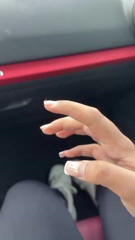 A close-up shot focuses on a light-skinned hand with long, square-shaped, light pink and white French tip nails. The hand is adorned with a large, ornate silver ring featuring a prominent square diamond surrounded by smaller diamonds on the ring finger. Initially, the hand is in a loose fist, then it extends two fingers into a peace sign, and finally, all fingers are extended, revealing the full set of nails and the ring. The person is wearing dark leggings, and light-colored shoes are visible on the floor below. The background shows the interior of a car, including a dashboard with a red trim accent. The video is filmed from a downward-looking, first-person perspective.