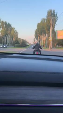 The video shows a person riding a motorcycle on a paved road. The person is wearing a black jacket and helmet. The motorcycle is red and black. The road is lined with trees on both sides. There are also streetlights and a pedestrian crossing sign visible. The sky is blue and clear, and the sun is setting, casting a warm light on the scene. The video is shot from inside a car, looking out through the windshield. The dashboard of the car is visible in the foreground.
