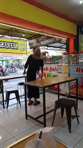 A person wearing a black dress and a hijab is standing in front of a counter. The person is bending down and appears to be cleaning a table with a cloth. There are tables and chairs set up in front of the counter. The counter has a sign that says "fresh juice". In the background, there are people walking by and motorcycles are being ridden. The environment appears to be an outdoor market or street vendor stall.