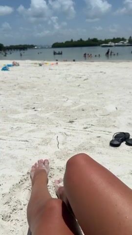 This video shows a first-person perspective of someone sitting on a sandy beach. Their legs and feet are in the foreground, with sand on their toes and legs. In the background, there is a body of water with several people swimming and playing. There are also some boats in the water, and a treeline with buildings in the distance. The sky is blue with scattered clouds. A pair of black flip-flops are visible on the sand to the right of the person's legs.