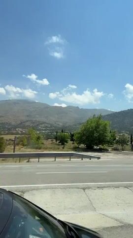 A landscape view from inside a vehicle, with the roof of the vehicle visible in the foreground. The video pans across a rural landscape under a bright blue sky with scattered clouds. Mountains dominate the background, with some appearing hazy. In the middle ground, there are fields, scattered trees, and some smaller hills covered in vegetation. A paved road with white and yellow lines runs horizontally across the frame. A single green tree stands prominently in the left-middle ground. The overall impression is a scenic drive through a hilly, possibly Mediterranean, region.
